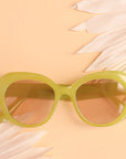 On a peachy background is a round pair of light green sunglasses with a light brown lens.