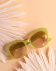 On a peachy background is a round pair of light green sunglasses with a light brown lens.