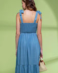On a green shadow background is a model wearing a blue flowy midi dress with tie shoulder straps and a square neckline.