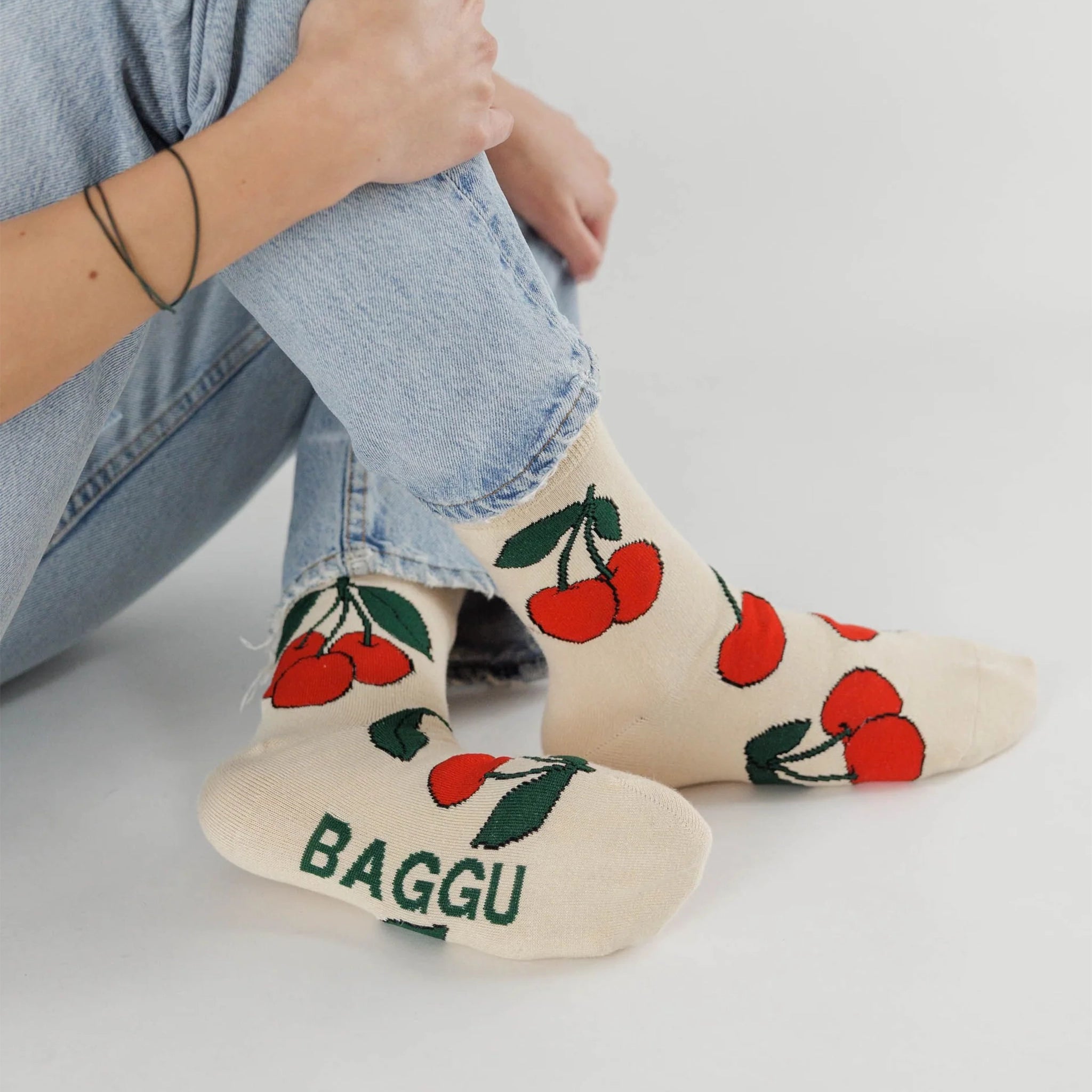 On a white background is a model wearing cream colored socks with a bright red cherries design on it along with &quot;Baggu&quot; on the bottom of the foot. 
