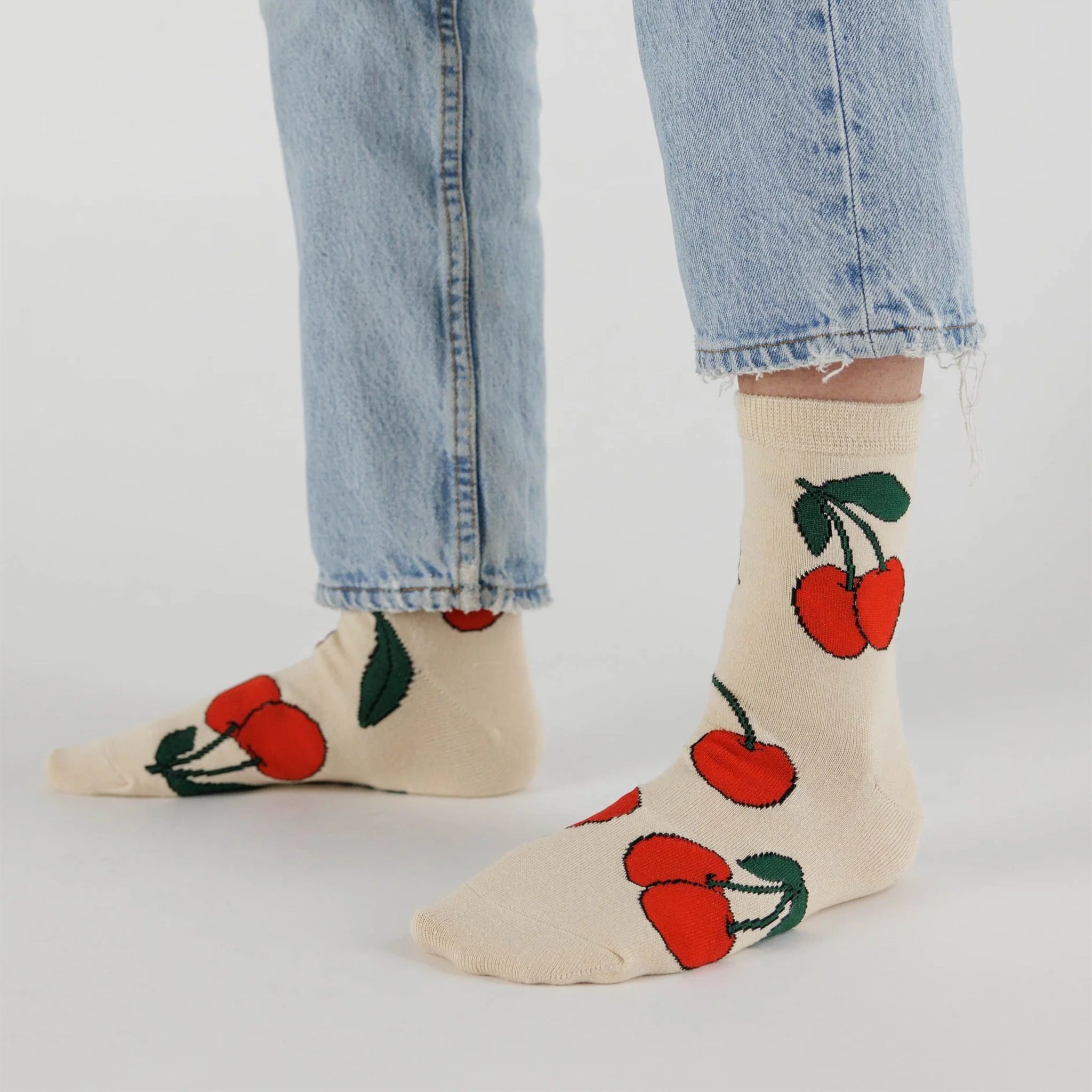 On a white background is a model wearing cream colored socks with a bright red cherries design on it along with "Baggu" on the bottom of the foot.