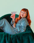 A person with long red hair lounges in a plush dark-green chair, wearing green-and-white striped pajamas and holding a pink cocktail glass while giving a playful wink.