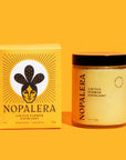 On a bright yellow background is a box with a design of a woman with cactus behind her head along with a jar of exfoliant that reads, "Nopalera Cactus Flower Exfoliant". They are both a similar shade of yellow and have a black screw on lid.