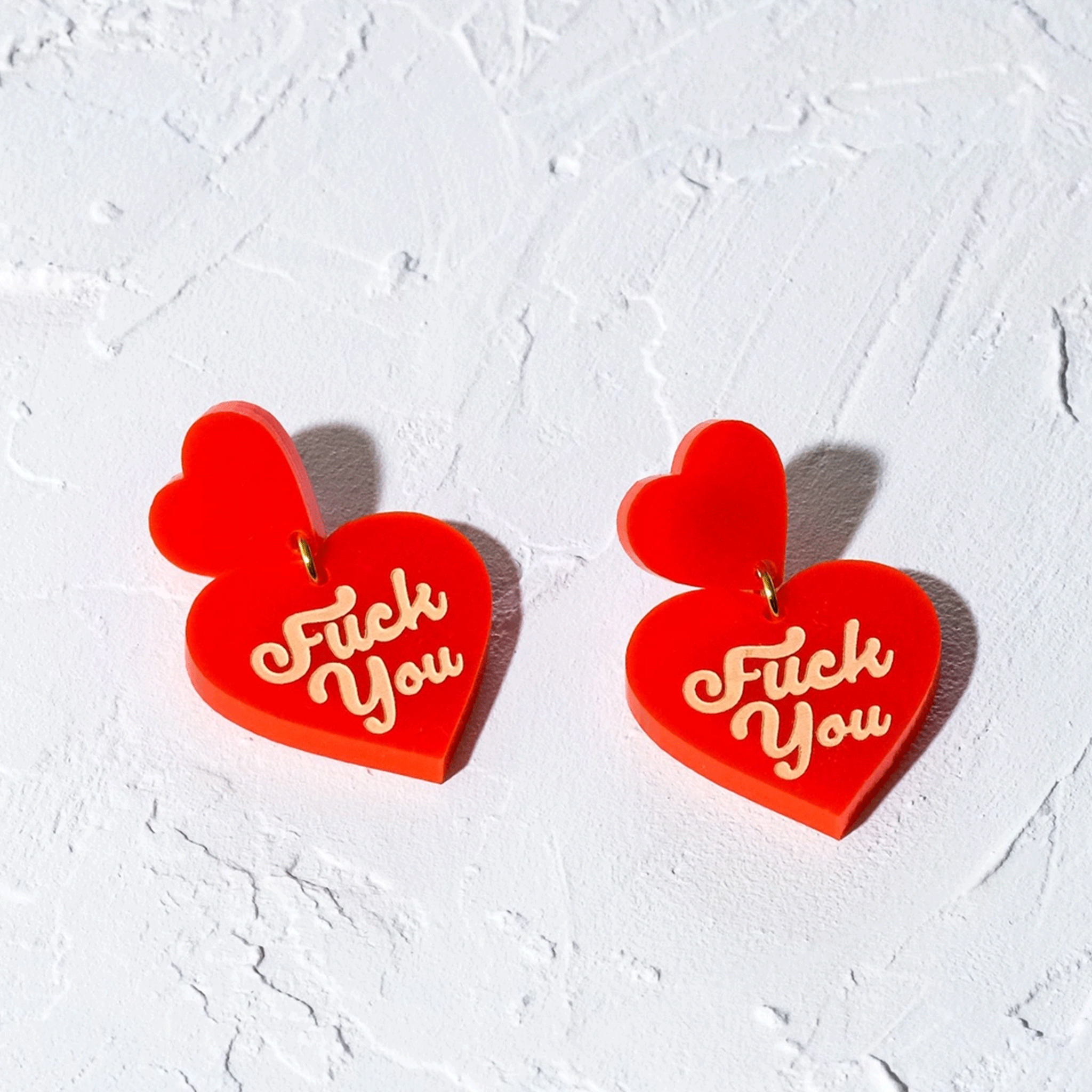 On a white background is a pair of heart double heart shaped earrings with text on each heart that reads, "Fuck You". 