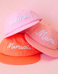 The three pink and orange flat brim hats with different white embroidery on the front reading, 'Cool Mom', 'Mama', and 'Mamacita' on the other.