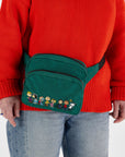 A dark green fanny pack with graphics of the characters of Peanuts and a black Baggu logo adjustable strap.