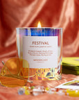 On a white background is a glass multicolored candle with a white label that reads, "Festival" and the scent notes.
