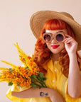 On an ivory background is a model wearing a pair of brown, flower shaped sunglasses and holding a bouquet of yellow flowers.