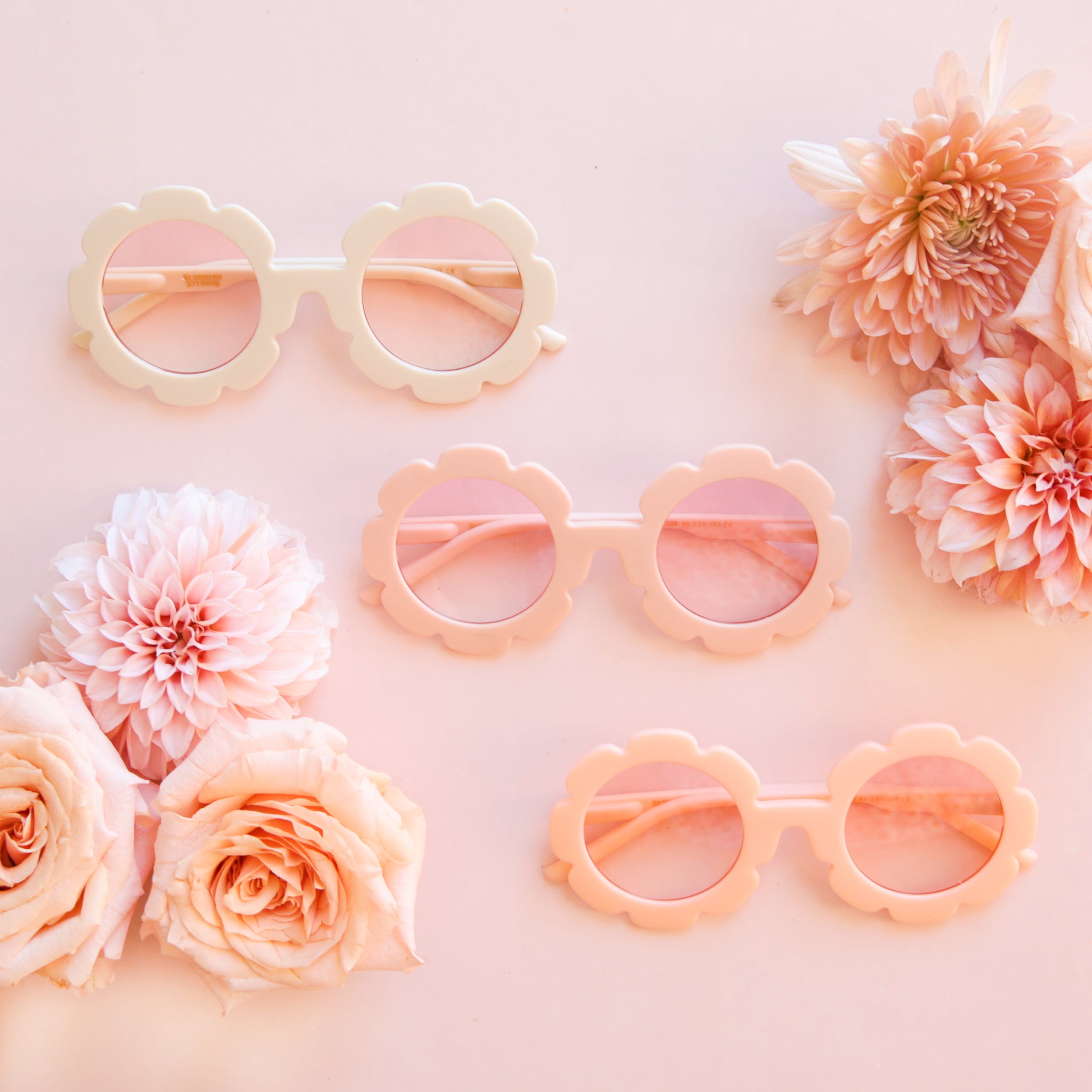 On a pink background is the three colors of flower shaped sunglasses surrounded with pink flowers. 