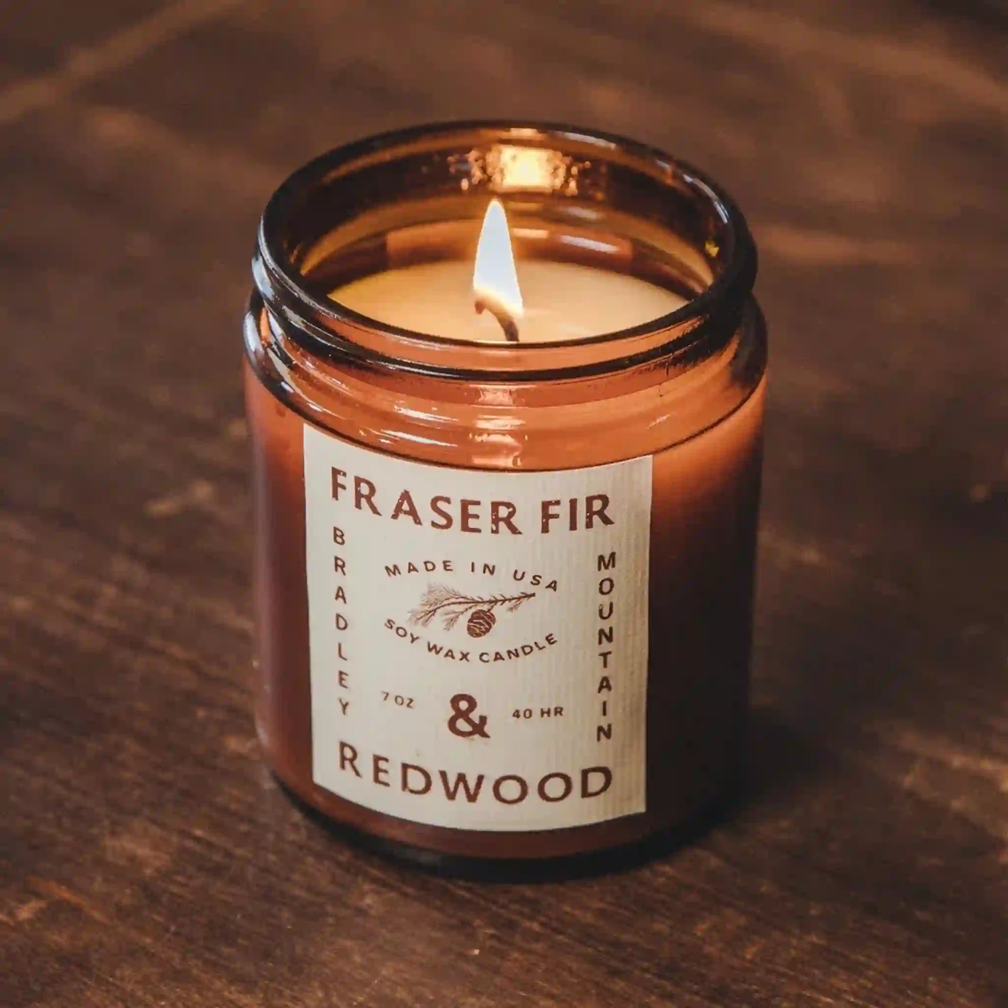 On a brown background is an amber brown glass candle jar with a tan label that reads, &quot;Fraser Fir &amp; Redwood&quot;. 