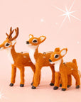 On a pink background is three different sized brown fur reindeer ornaments. There is a baby, a mama and a dad with antlers. Each ornament sold separately.