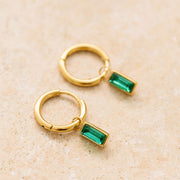 On a tan background is gold hoop earrings with an emerald cut emerald stone hanging from it.