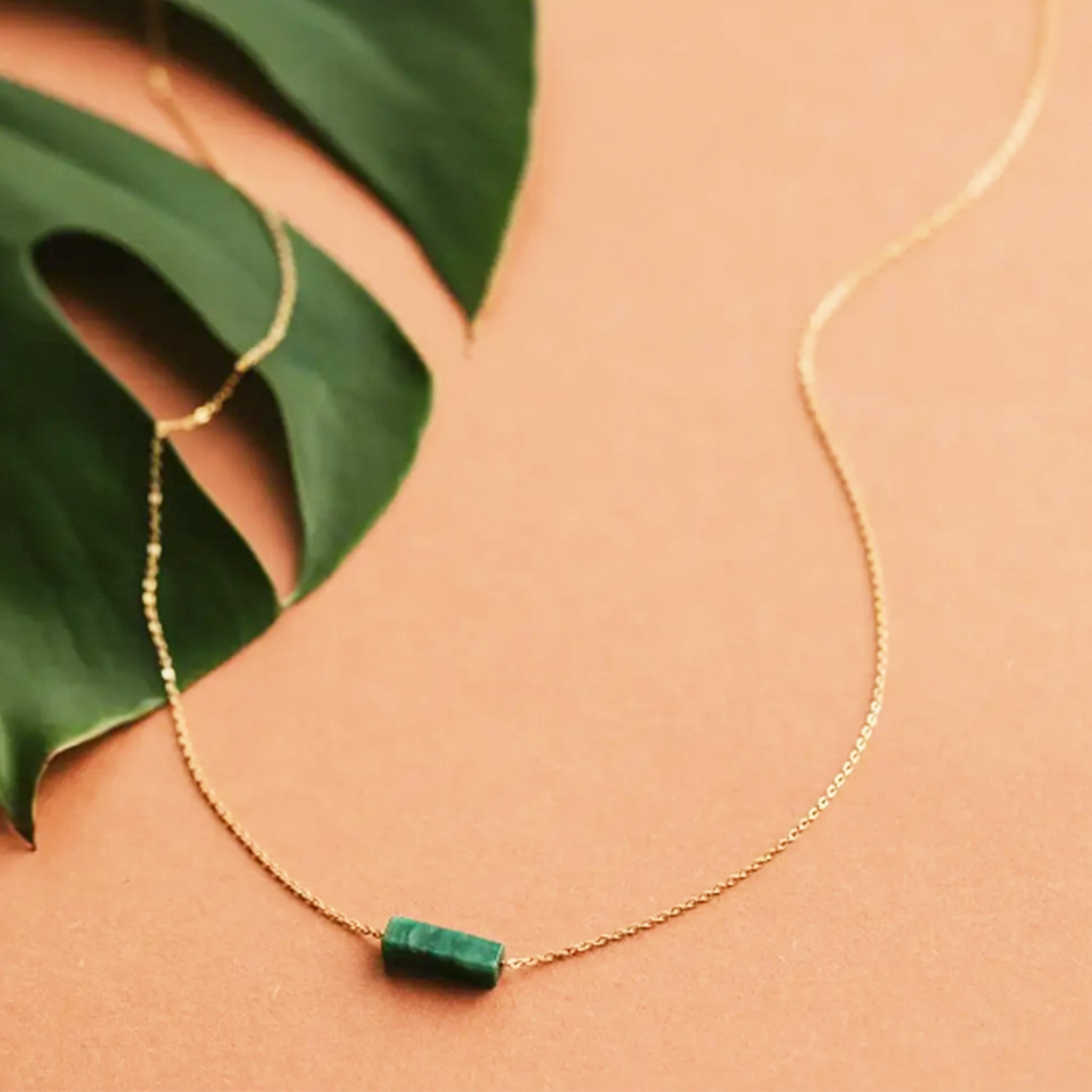 On a neutral background is a gold chain necklace with a rectangle green stone in the center. 