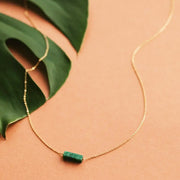 On a neutral background is a gold chain necklace with a rectangle green stone in the center.