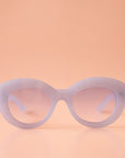 On a peachy background is a pair of light blue sunglasses with a rounded shape and a slight cat eye corner. The lenses are also a light blue shade