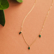 On a neutral background is a gold chain necklace with three rectangle emerald stones dangling from three different points on the chain.