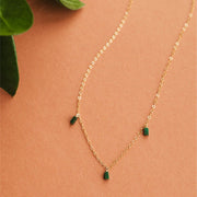 On a neutral background is a gold chain necklace with three rectangle emerald stones dangling from three different points on the chain. 