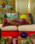 Long red velvet cushion with a colorful floral print and olive green tassels is shown on a couch.