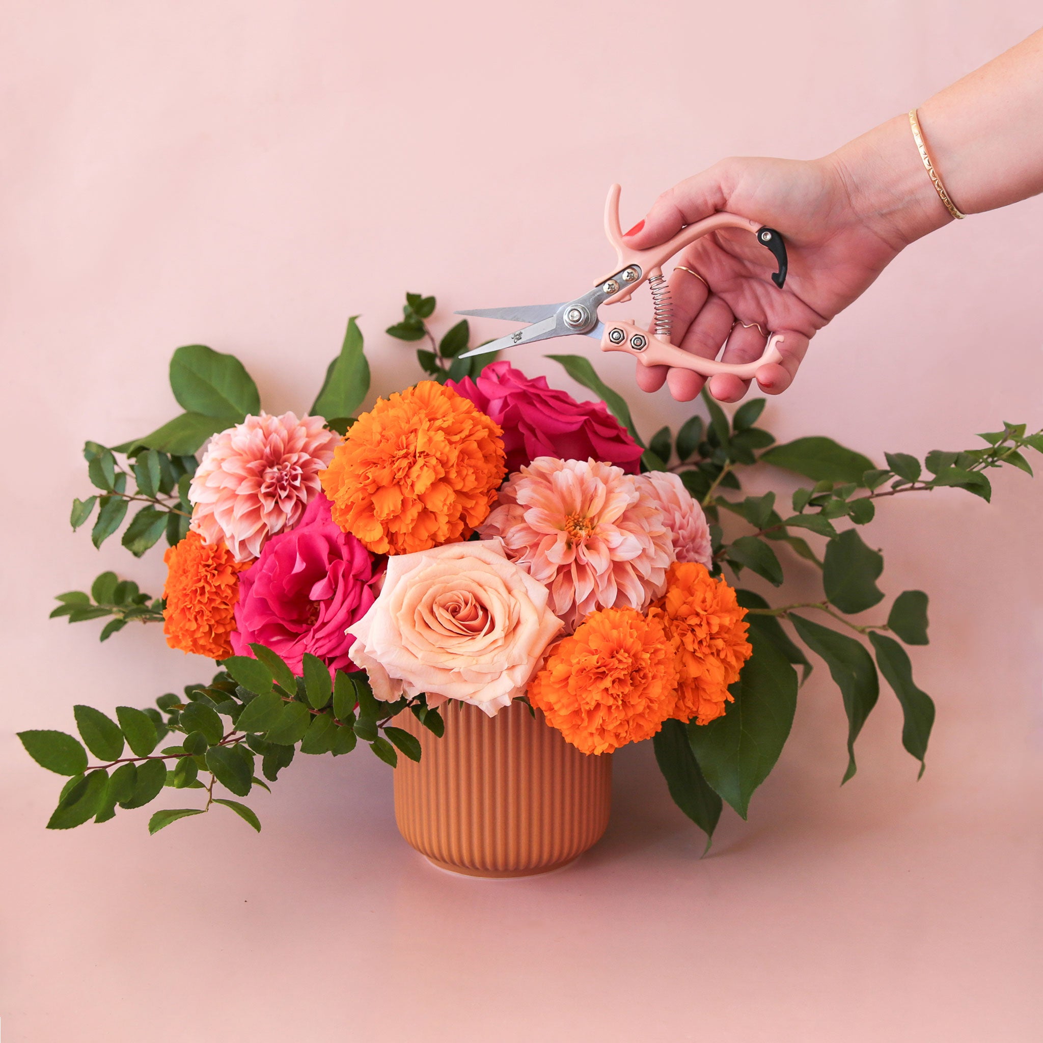 a floral arrangement with flowers of pink and orange sits in a terracotta fluted pot with a handing reaching in holding a pair of pruning shears