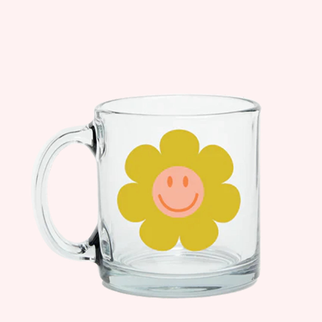 On a light pink background is a clear glass mug with an yellow smiley face daisy graphic in the center. 