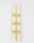 On a white background is a pair of ivory taper candles with a gold bow design.