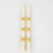 On a white background is a pair of ivory taper candles with a gold bow design. 