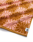 A pink and burnt orange bath/beach towel with a wavy floral checker design in each of the squares photographed here laid flat on a white background.