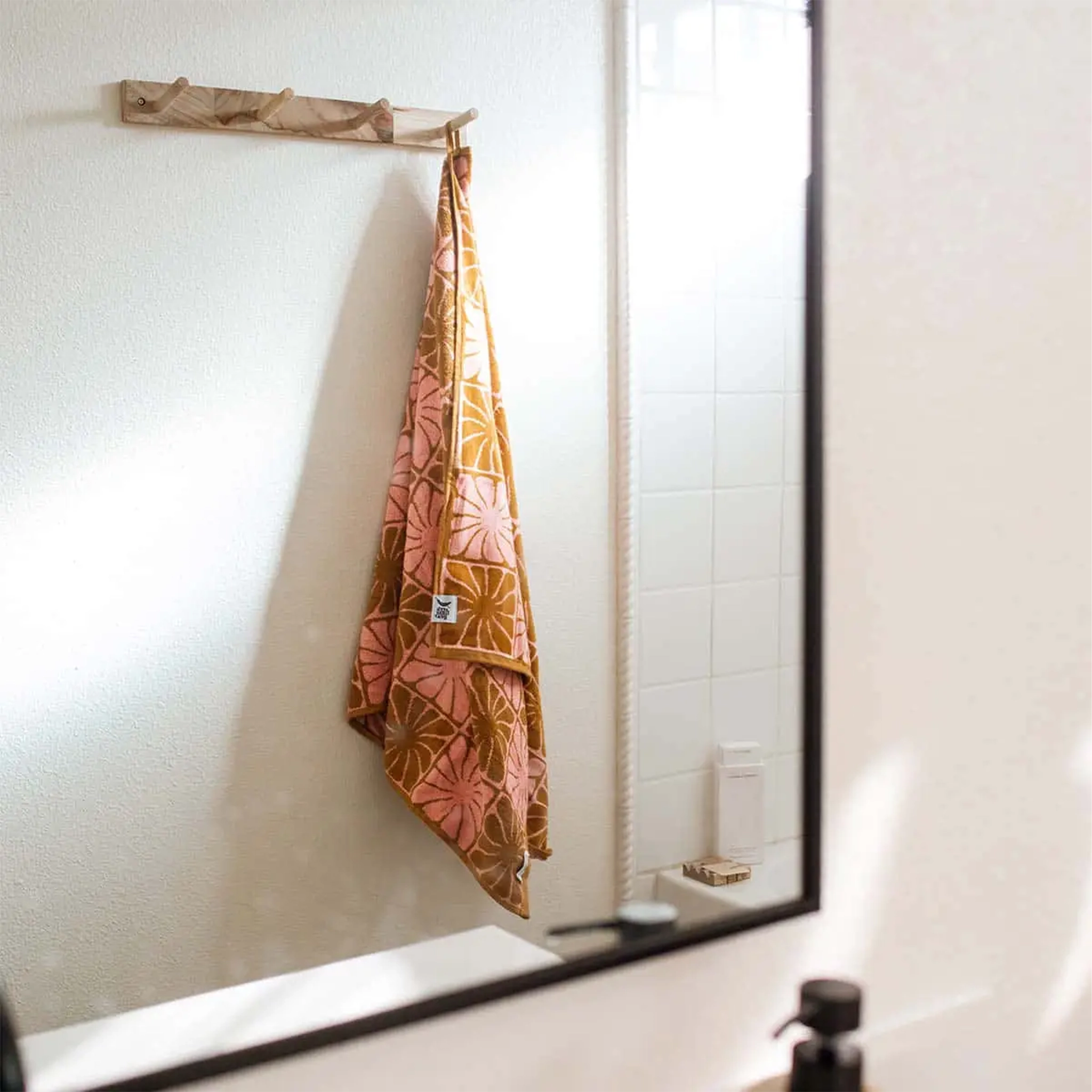 A pink and burnt orange bath/beach towel with a wavy floral checker design in each of the squares photographed here hung up on hooks in a bathroom. 