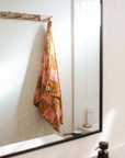 A pink and burnt orange bath/beach towel with a wavy floral checker design in each of the squares photographed here hung up on hooks in a bathroom.