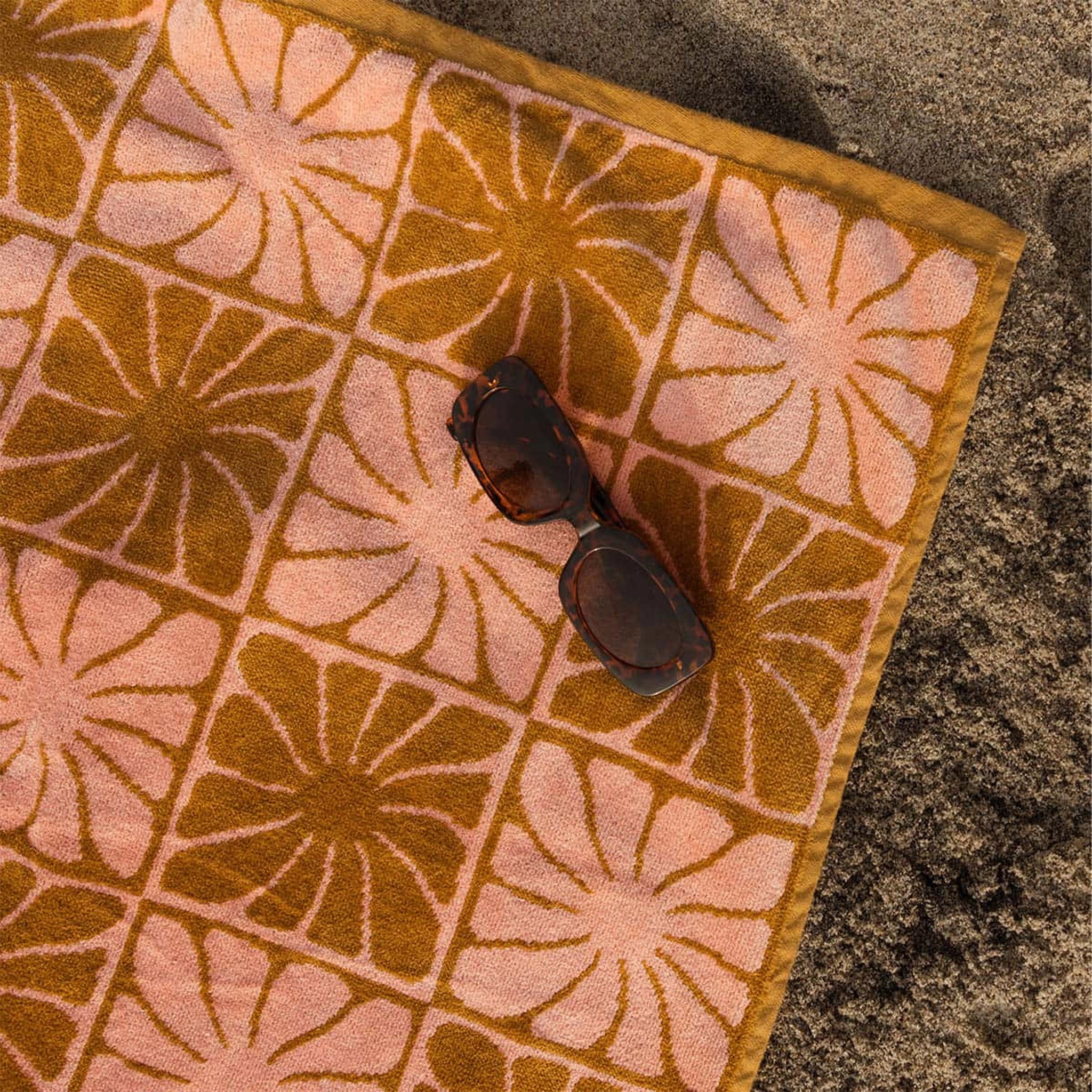 A pink and burnt orange bath/beach towel with a wavy floral checker design in each of the squares photographed here on a sandy surface staged next to a pair of sunglasses. 