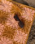A pink and burnt orange bath/beach towel with a wavy floral checker design in each of the squares photographed here on a sandy surface staged next to a pair of sunglasses.