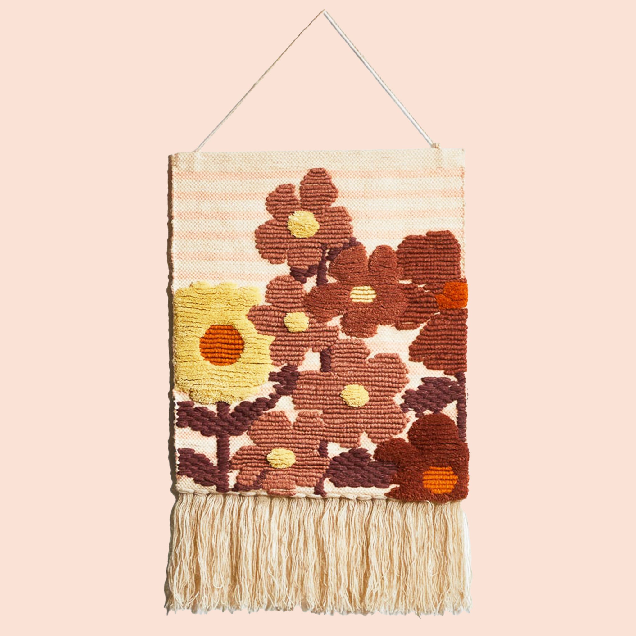 A hanging piece of woven wall art featuring rust and yellow colored floral prints and a fringe detail hanging from the bottom edge.