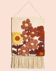 A hanging piece of woven wall art featuring rust and yellow colored floral prints and a fringe detail hanging from the bottom edge.