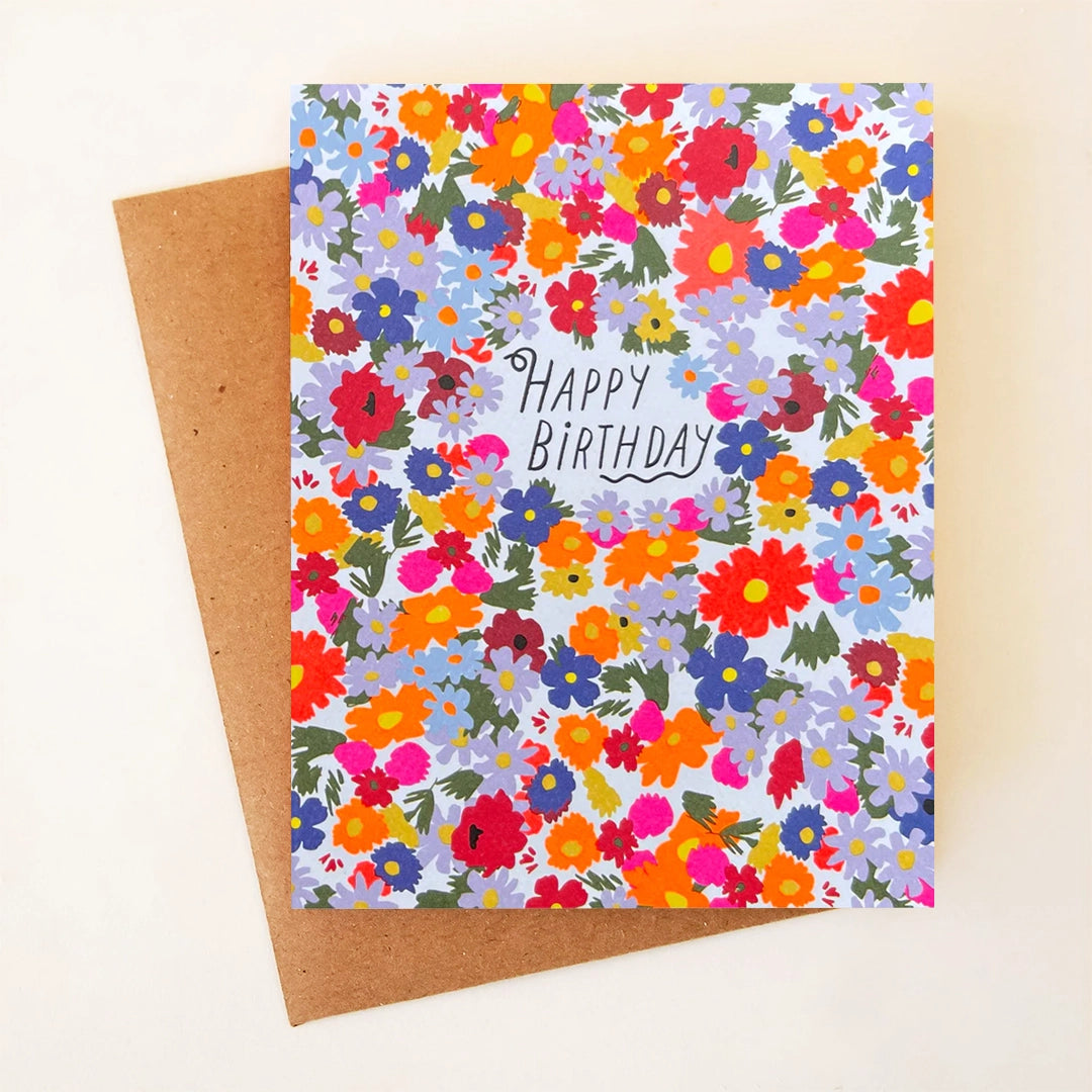 On a cream background is a greeting card with a multicolored floral print with text in the center that reads, &quot;Happy Birthday&quot; along with a kraft envelope. 