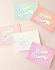 On a tan background is five greeting cards in different colors that read, "Happy Holidays" with holographic rays coming out from the text in the center. This pack of card includes one pink, white, light pink, peach and teal card.
