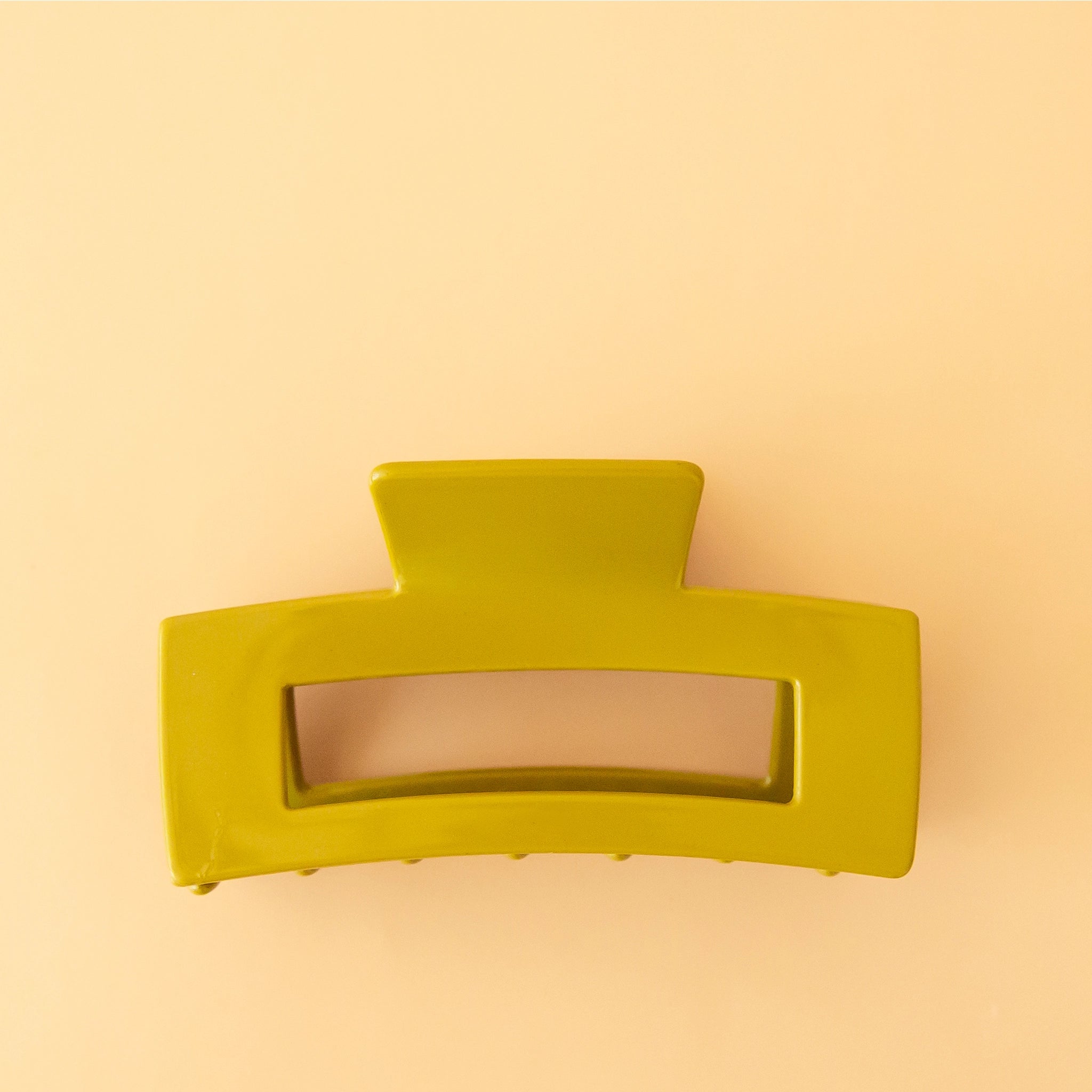 On a peachy background is the Havana Hair Claw in the shade Avocado which is a green rectangle shaped hair clip.