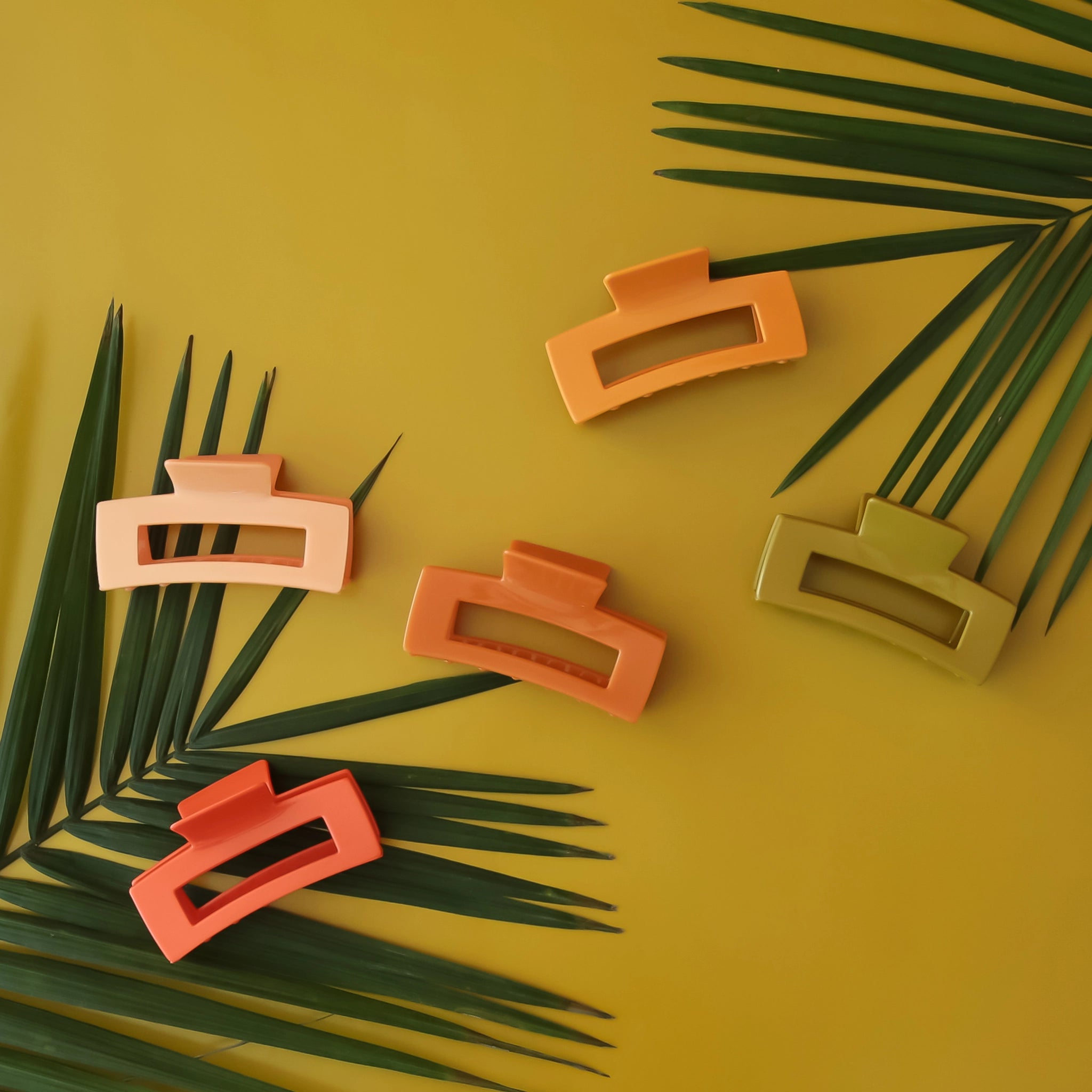 On a green background is a variety of different colored hair clips that have a rectangular design. The colors included are, red, dark orange, yellow/orange, light orange, and olive green. 