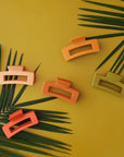 On a green background is a variety of different colored hair clips that have a rectangular design. The colors included are, red, dark orange, yellow/orange, light orange, and olive green.