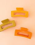 On a peachy background is three rectangle claw clips in different colors. One yellow, one olive, and one orange.