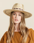 On a neutral background is a model wearing a tan woven sun hat with a wide brim and brown stitching details around the bottom of the brim as well as around the base of the hat.
