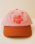 A light pink and orange baseball cap with an orange embroidered hibiscus flower in the center.