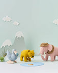On a light blue background is a set of extinct stuffed animals. A blue dodo bird, a yellow sabre tooth tiger, and a pink mammoth.