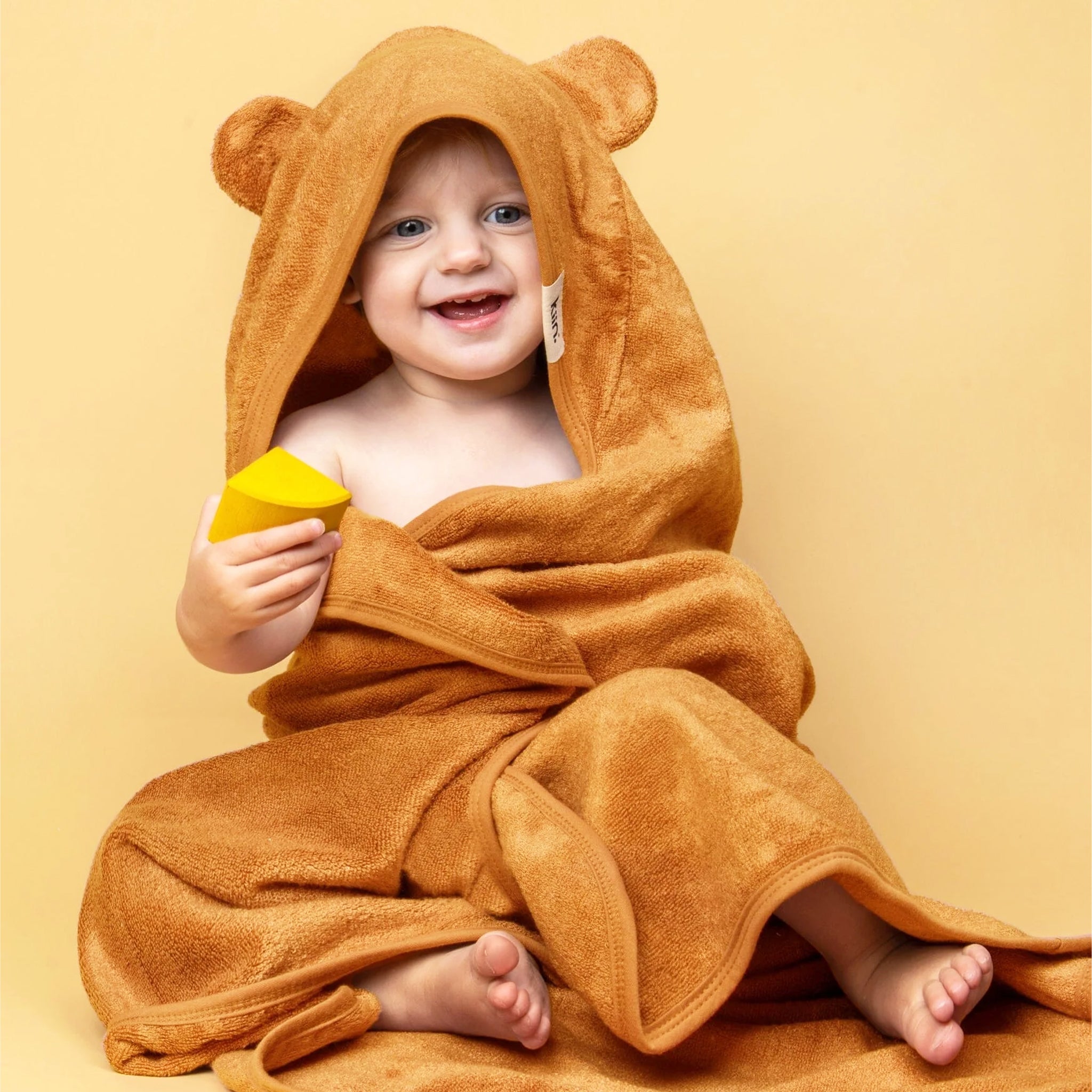 On a yellow background is a caramel orange colored bath towel for kids with a hood and bear ears on the hood. 