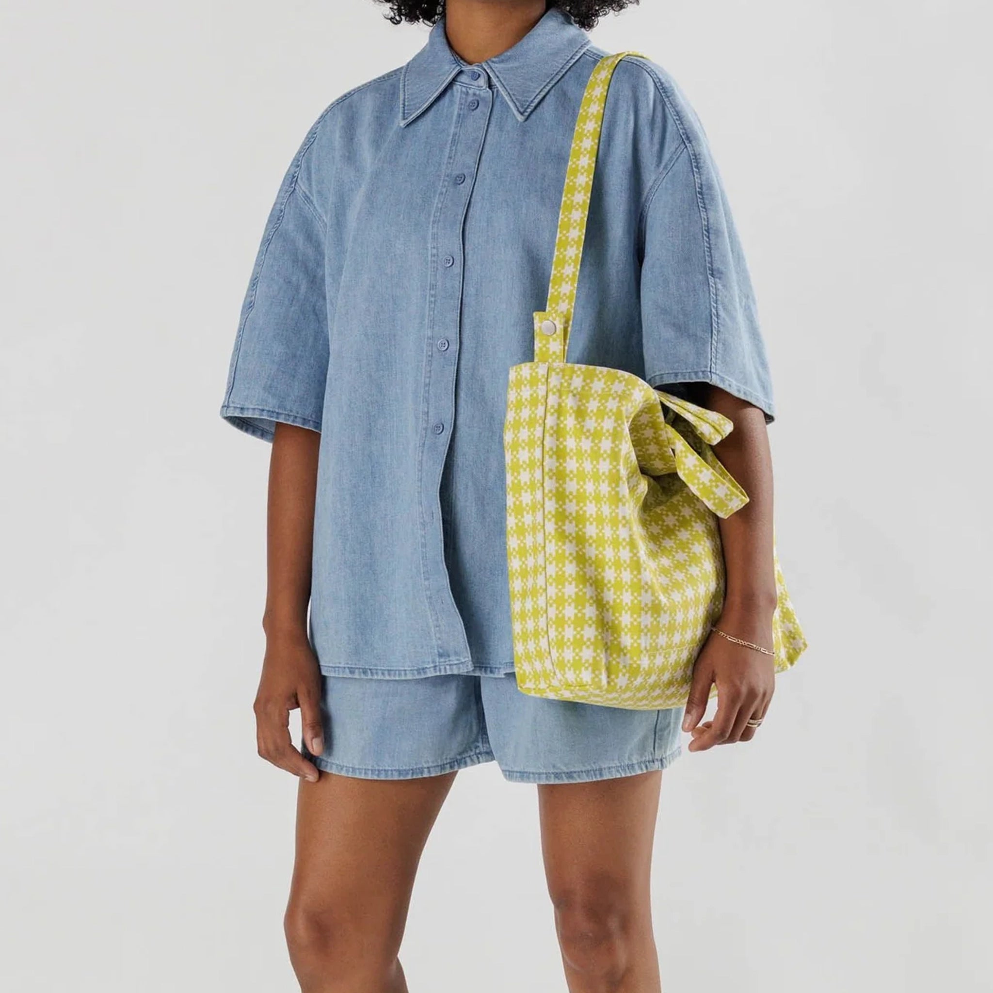 On a grey background is a model wearing a neon green pixel gingham print tote bag with a shoulder strap and hand straps .