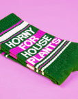 On a purple background is a green and bright pink pair of socks with white text that reads, "Horny For House Plants"