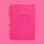 On a pink background is a hot pink spiral bound notebook with a soft cover and text in the center that reads, "Howdy" along with a pink and white horseshoe print on the inside cover.