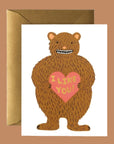 A white folded card with a brown bear graphic on the front holding a pink heart that says, "I Like You" in black writing.