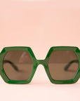 On a peachy background is a pair of hexagon shaped plastic sunglasses in an emerald green color with a dark brown lens.