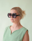 On a mint green background is a model wearing the Iris sunglasses in the shade black.