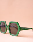 On a peachy background is a pair of hexagon shaped plastic sunglasses in an emerald green color with a dark brown lens.
